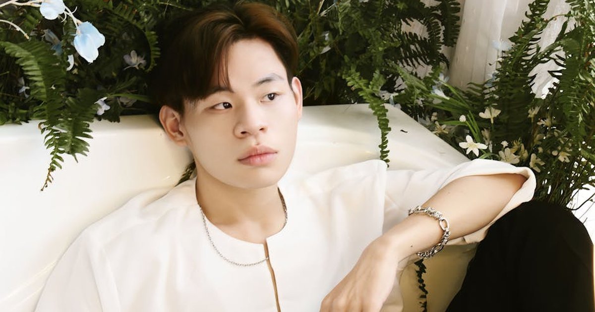 Asian man sitting elegantly on a sofa surrounded by lush greenery. Wearing jewelry.