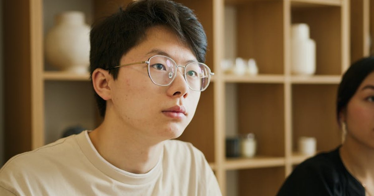 Portrait of a young man wearing eyeglasses, seated indoors with a blurred background.