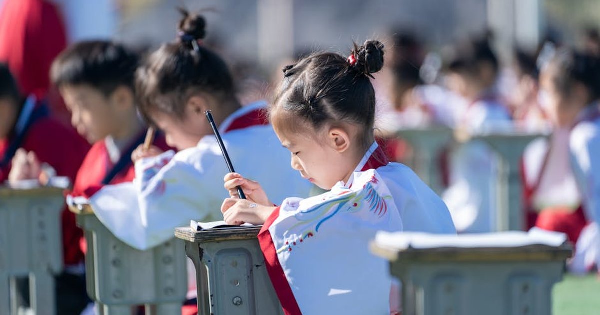 A young girl in traditional dress writes outdoors on a sunny day, focusing intently.