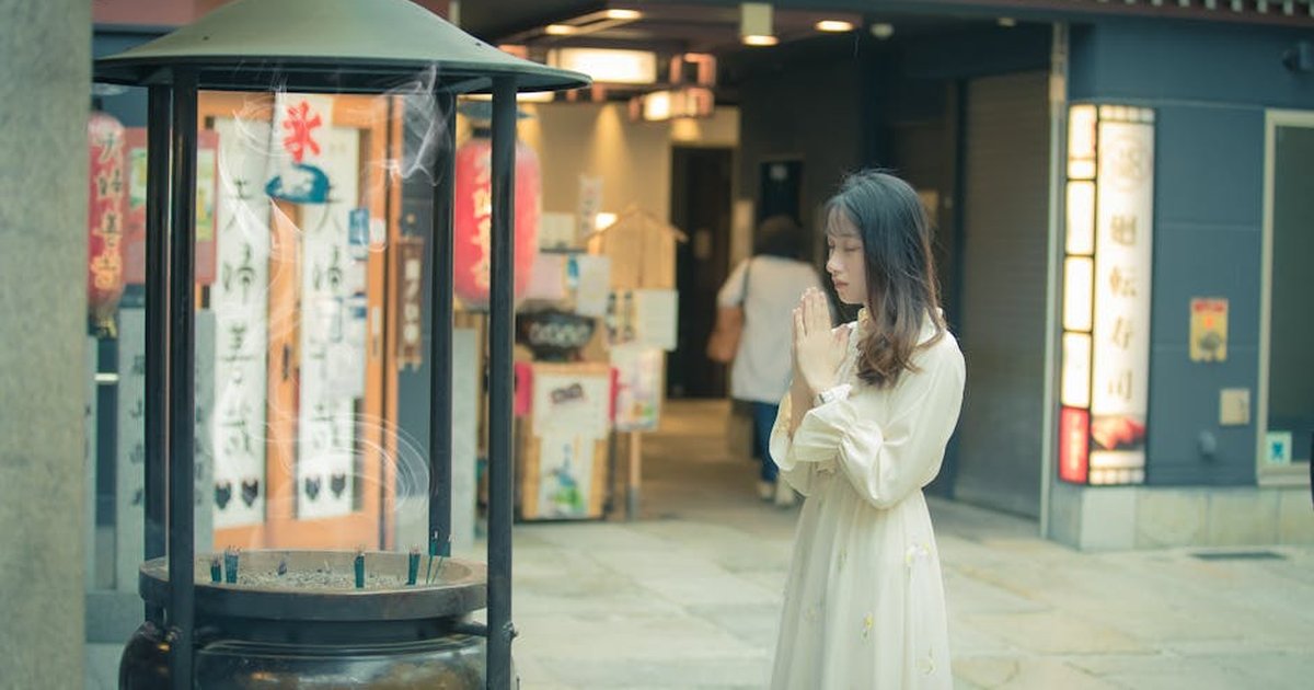 A woman in a white dress praying beside an incense burner in a serene temple setting.