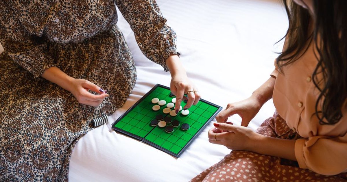 High angle crop faceless females in stylish dresses sitting on comfy bed and playing Othello board game