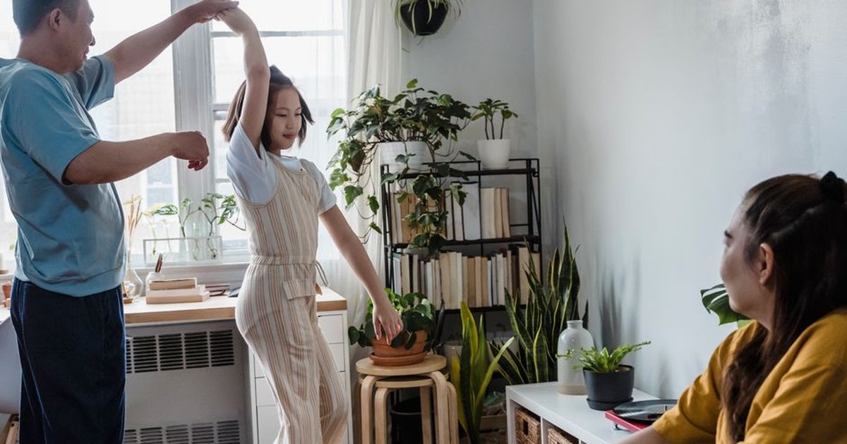Father dancing with daughter in a sunlit room as mother watches, surrounded by indoor plants.