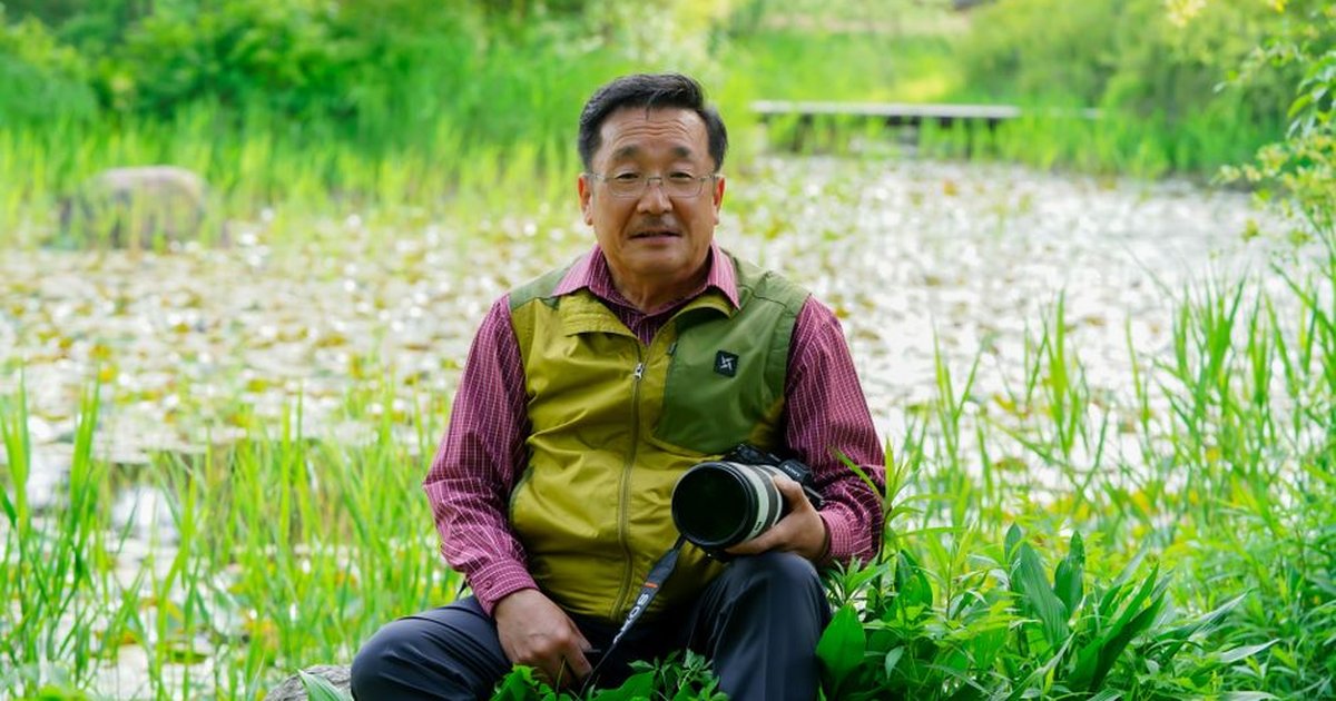 An Asian man sitting by a pond, holding a camera amidst lush greenery.