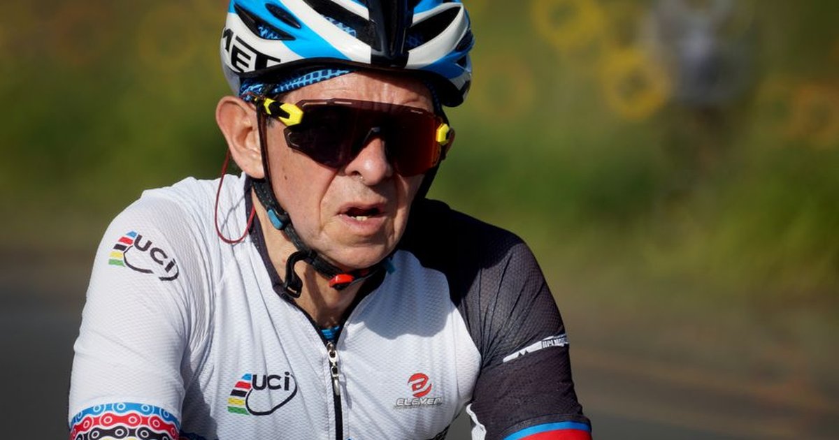 Elderly man cycling in a competitive outdoor race wearing helmet and sunglasses.