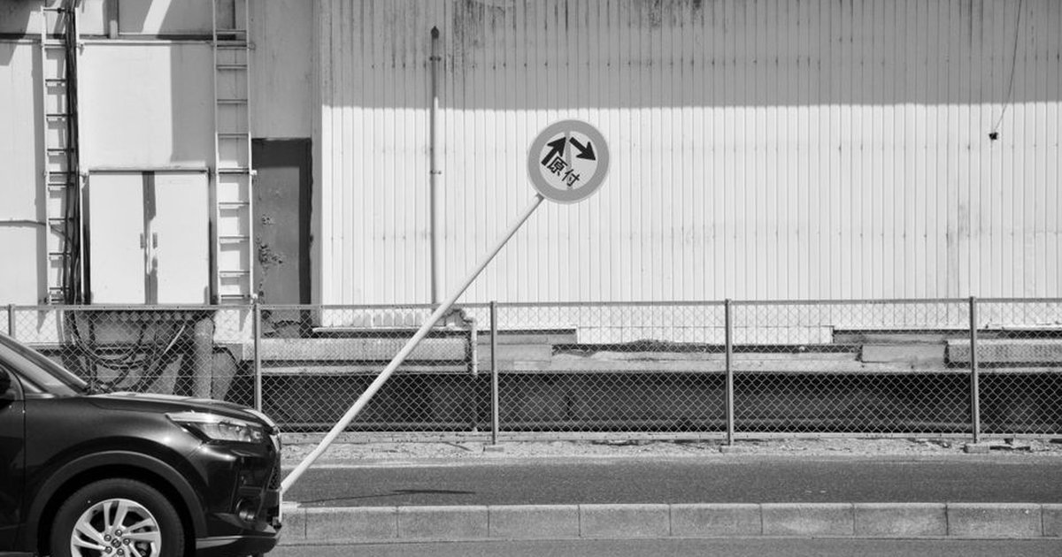 Black and white image of a leaning traffic sign beside a parked car and industrial wall.