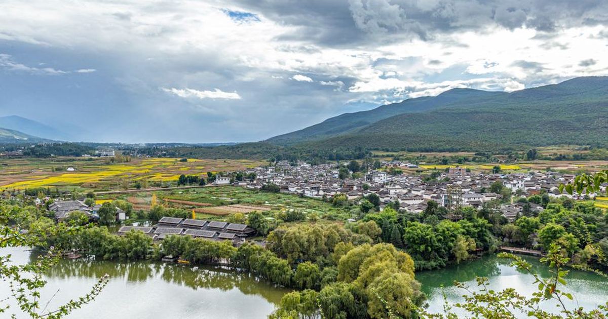 Picturesque view of a mountain town with lush fields and a serene lake under dramatic skies.