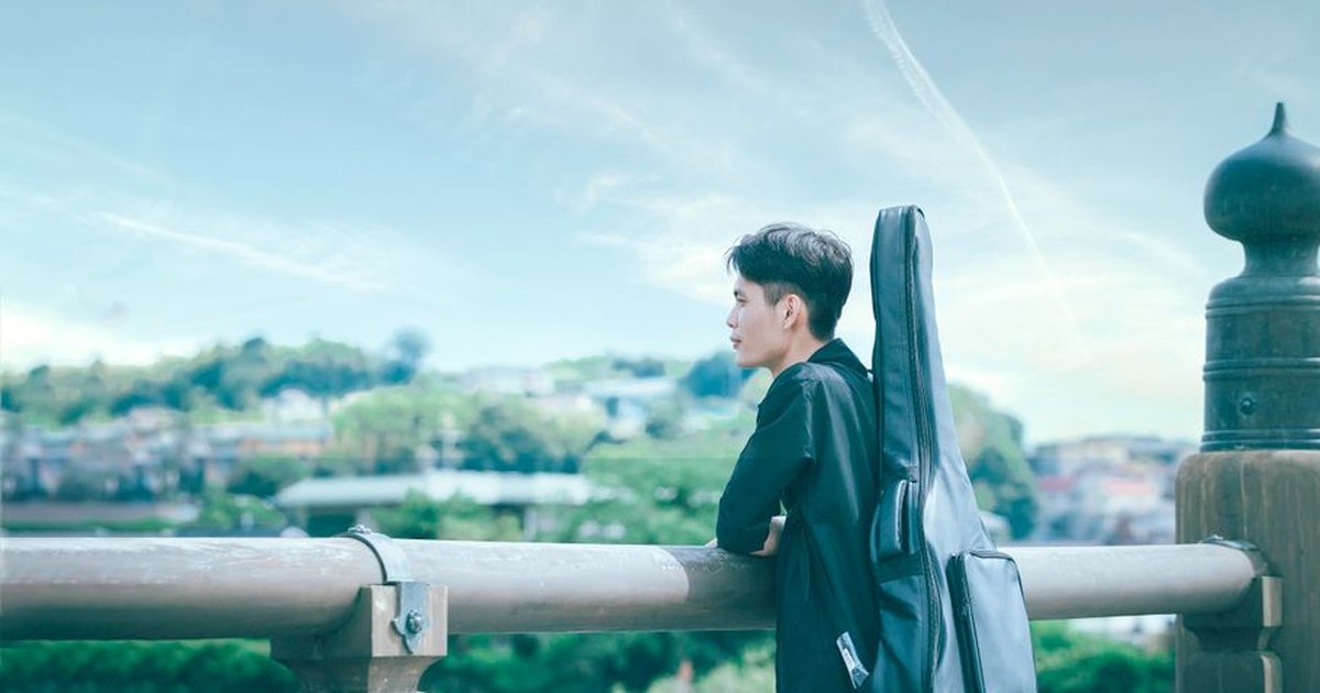 A young man with a guitar case standing on a bridge, overlooking a scenic landscape.