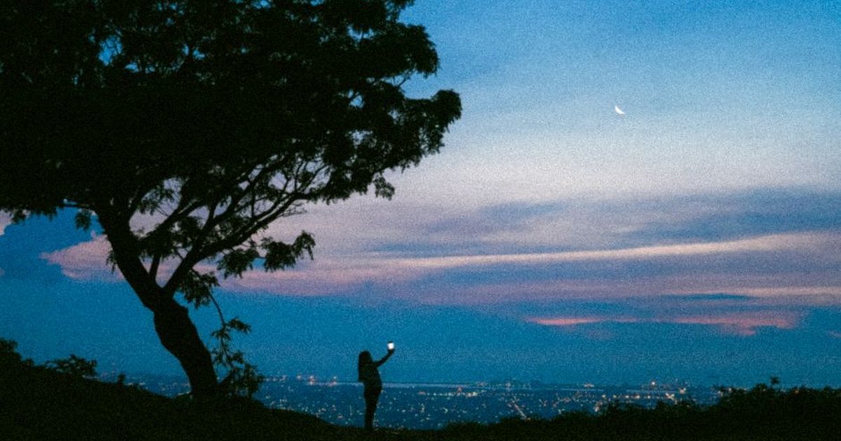 A silhouette of a person taking a photo against a cityscape during dusk with a visible crescent moon.
