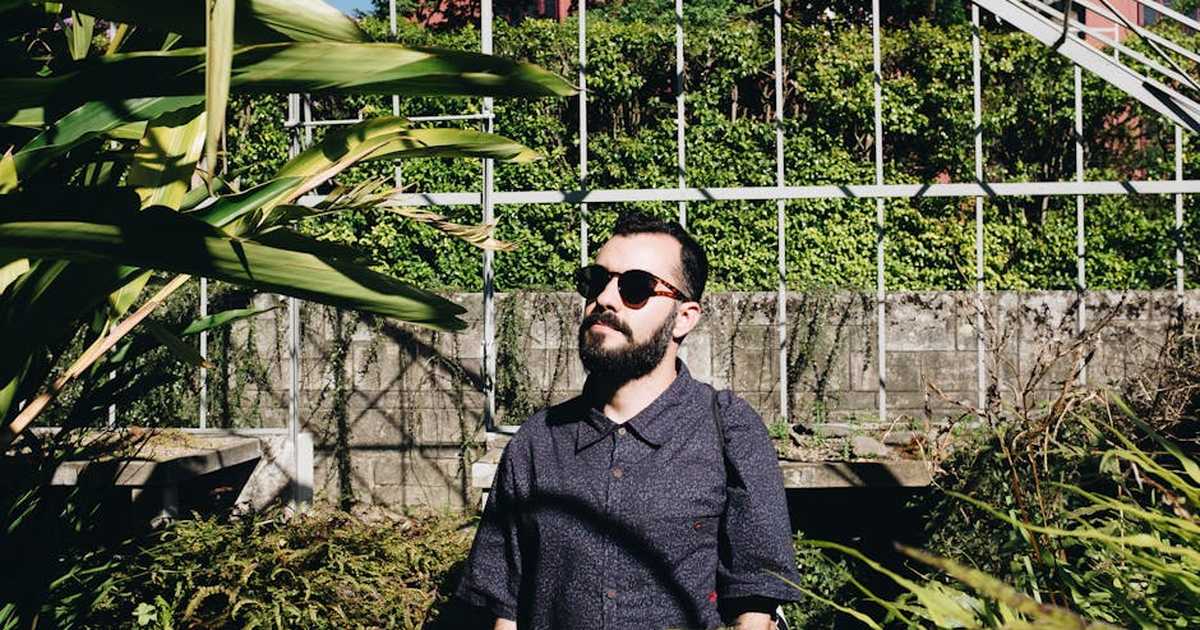 A bearded man wearing sunglasses stands in lush green surroundings and sunlight outdoors.