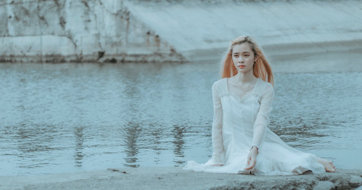 Calm young Asian female in elegant white dress sitting on stone surface near water in daytime