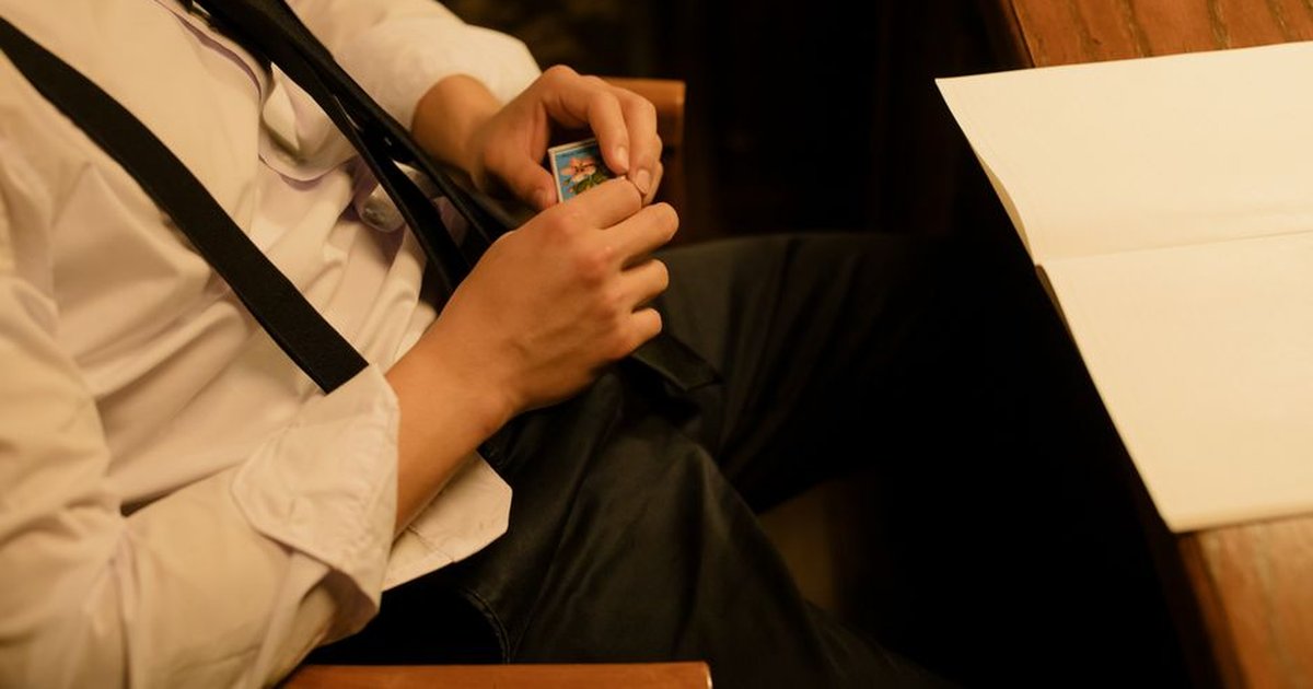 A man in suspenders holds a matchbox while sitting at a wooden desk in a dimly lit room.