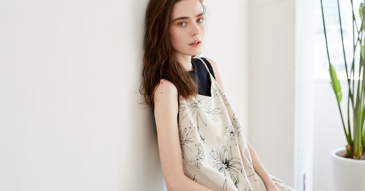Caucasian woman in a floral dress leans against a white wall with indoor plants nearby.
