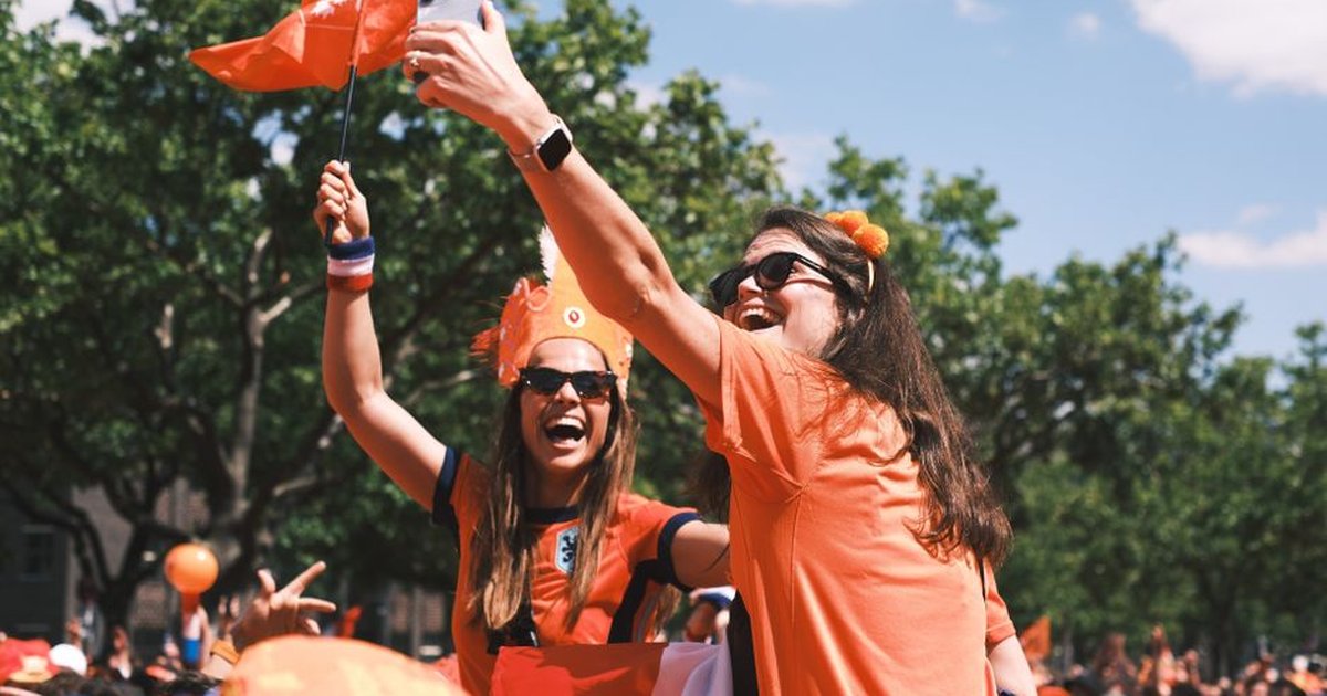 Excited Dutch football fans take selfies and celebrate outdoors wearing orange, waving flags.