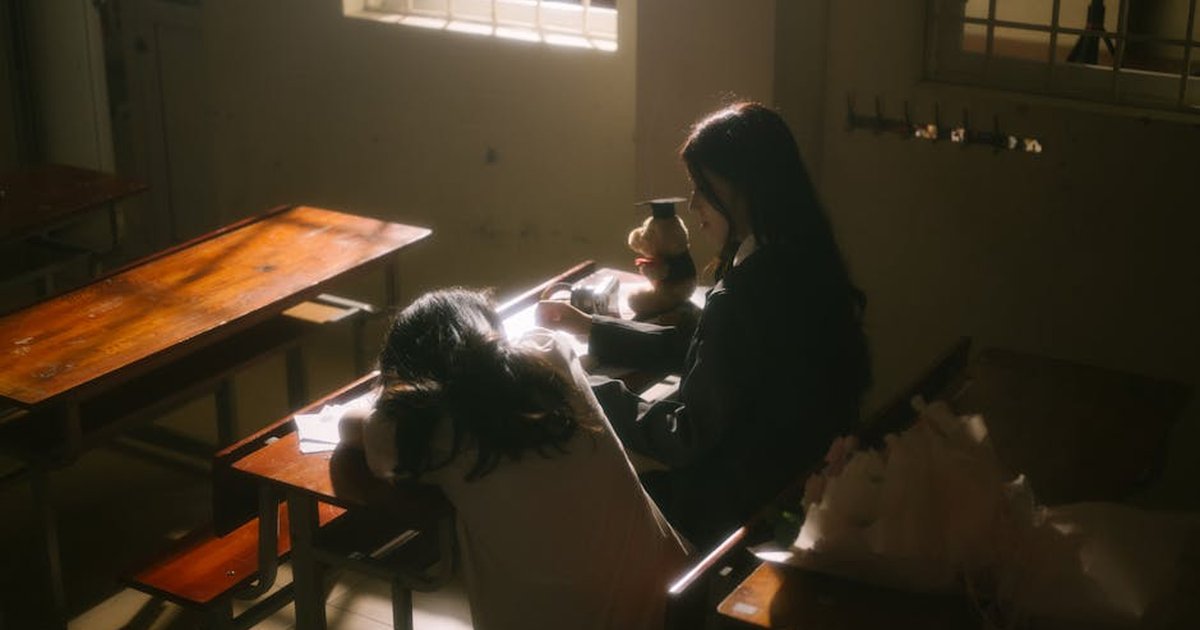 Two students in a dimly lit classroom, one resting on a desk, creating a moody atmosphere.