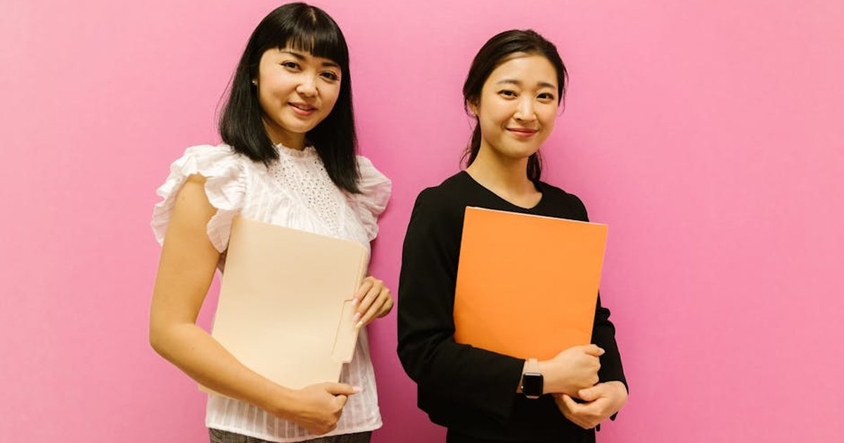 Two professional women smiling and holding folders against a pink background. Ideal for business and corporate themes.