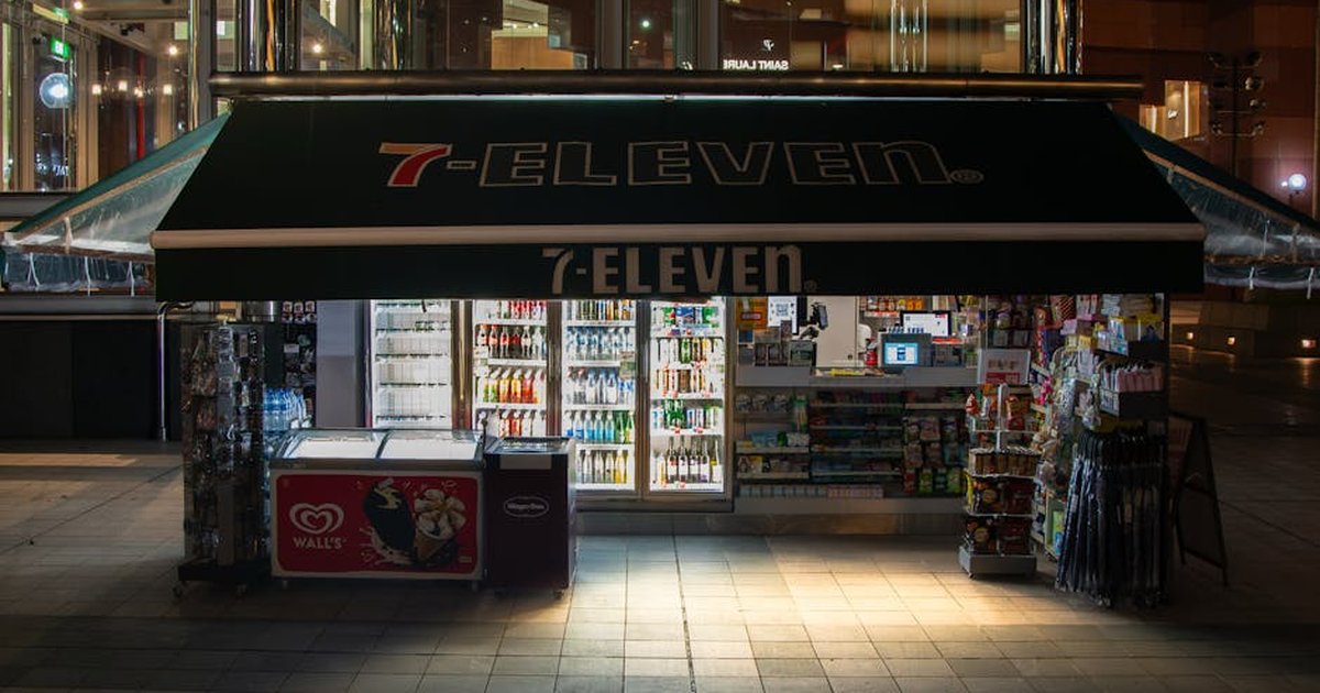 A brightly lit 7-Eleven convenience store at night, showcasing drinks and snacks.