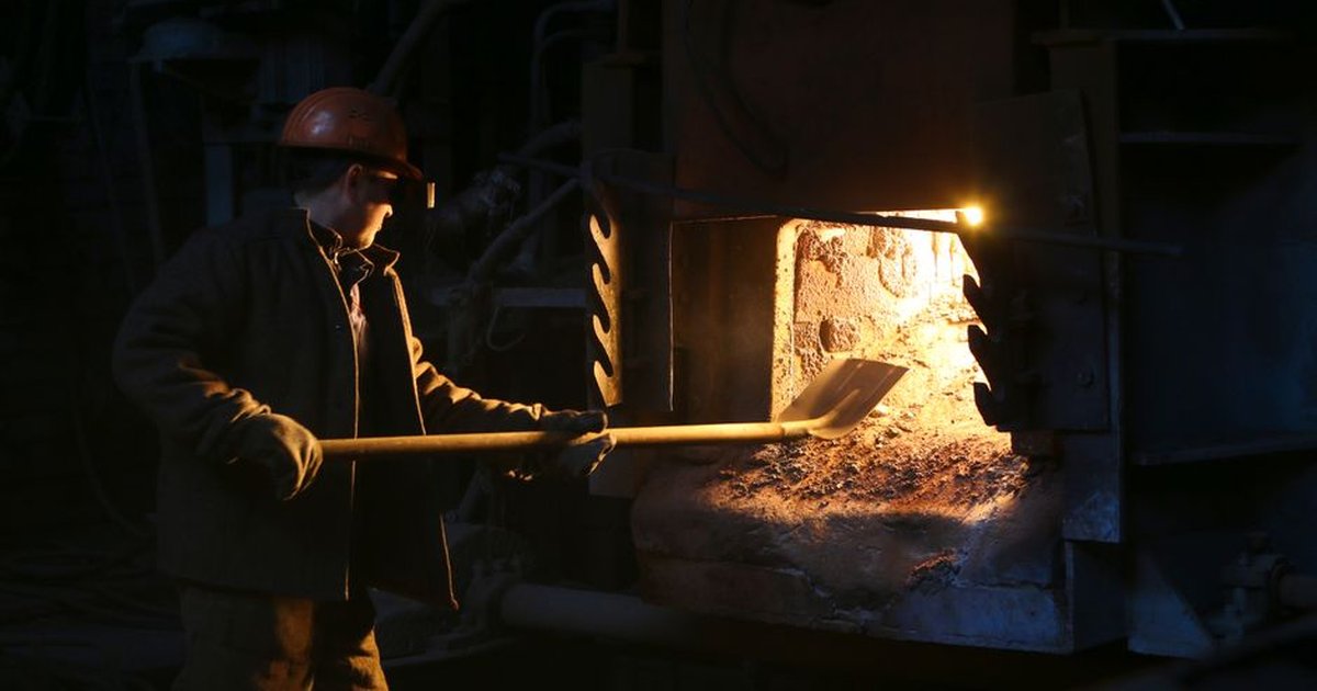 A man in a hard hat works with a furnace using a shovel, showcasing industrial labor.