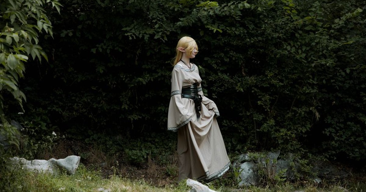 A cosplayer dressed as an elf in a fantasy costume stands in a lush, green forest.