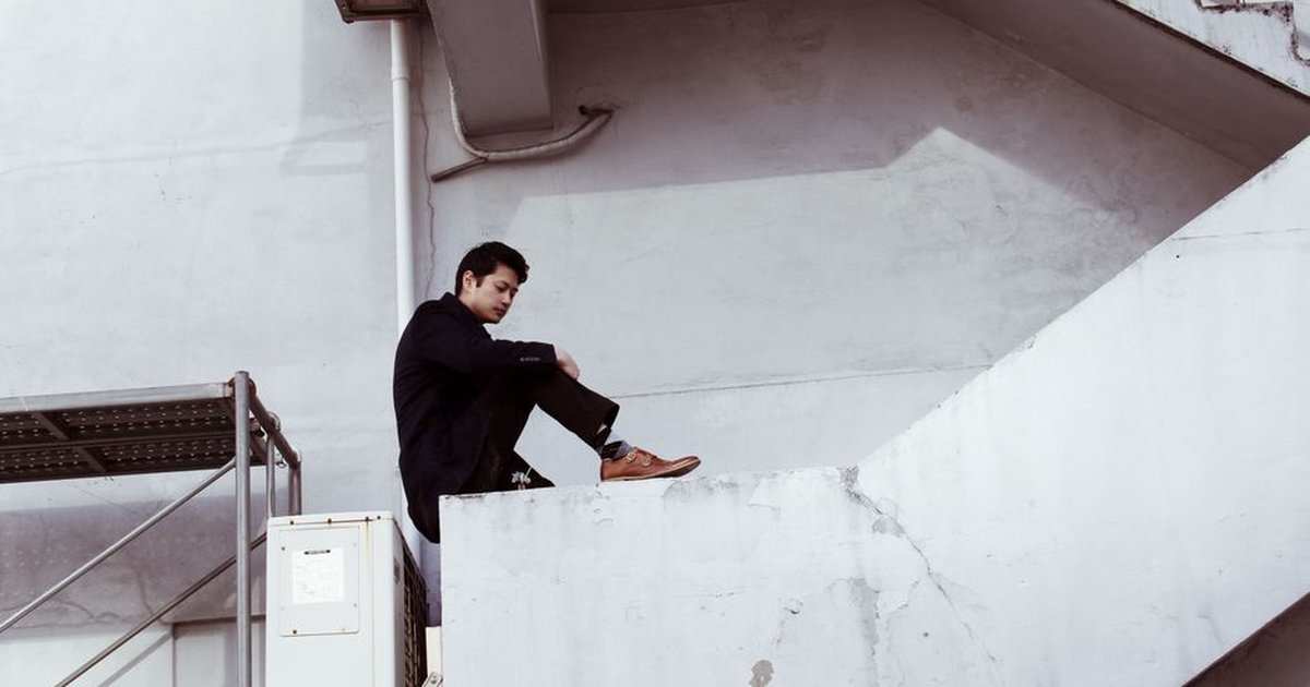 Asian man in a black suit sitting on concrete stairs, showcasing fashion and architecture.