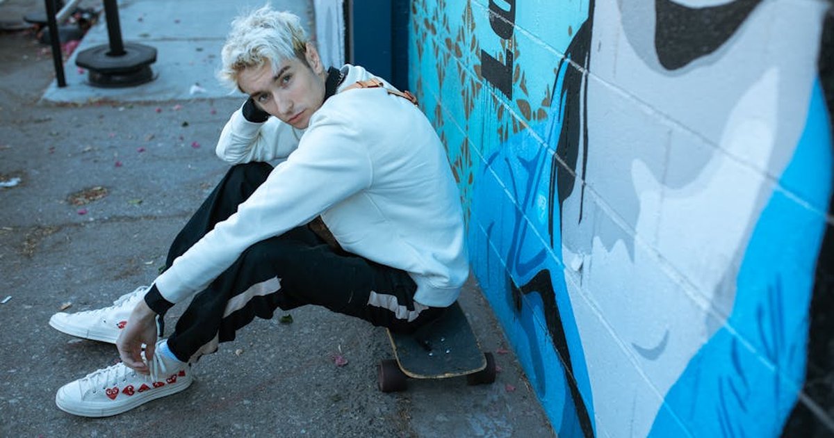 A fashionable young man sits on a skateboard next to graffiti art on an urban street.