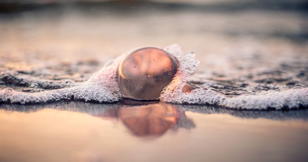 A glass ball reflects the ocean sunset as waves crash around it on a beach in Świnoujście.