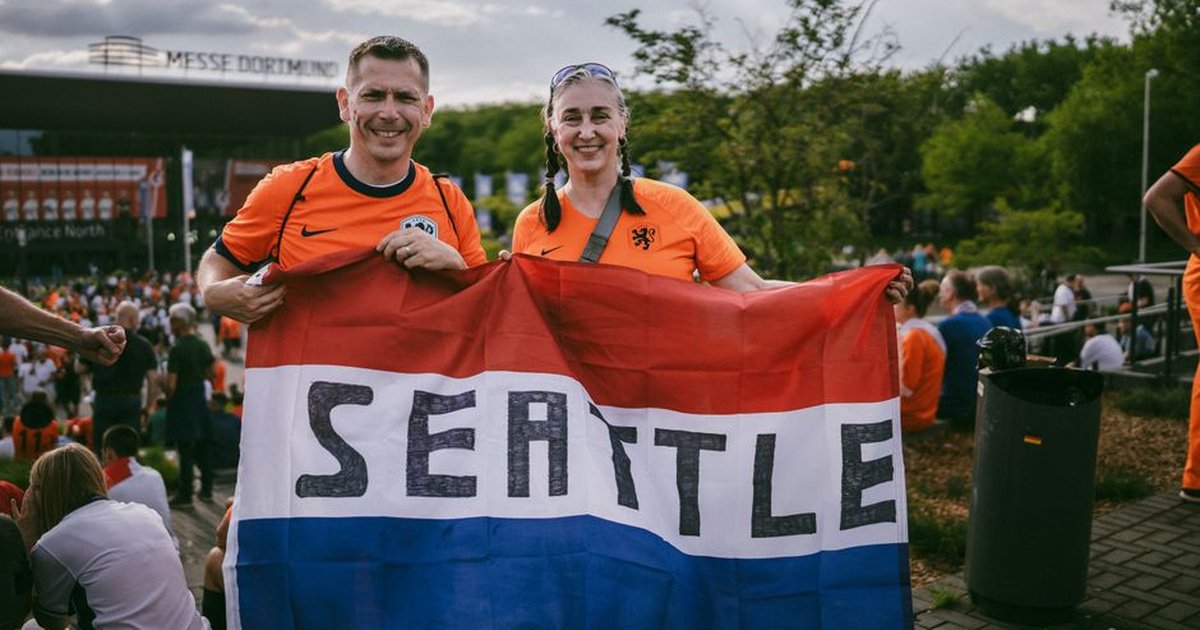 Two soccer fans in orange jerseys holding a Seattle flag outside a stadium in Dortmund.