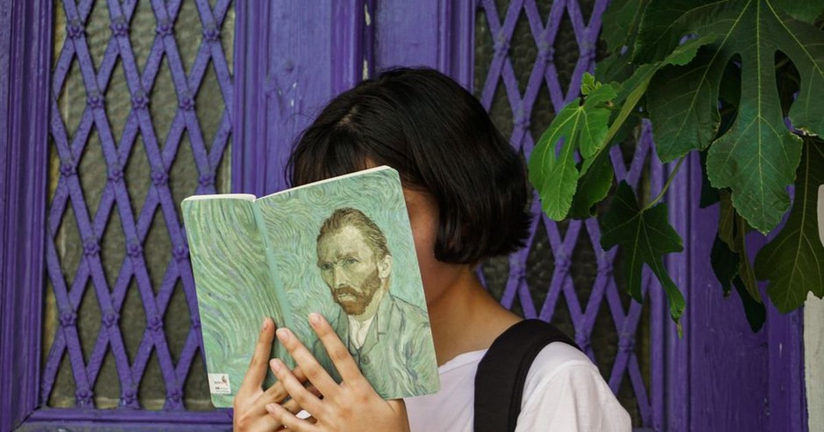 A young woman reads a Van Gogh art book in front of a purple door, partially hidden by fig leaves.