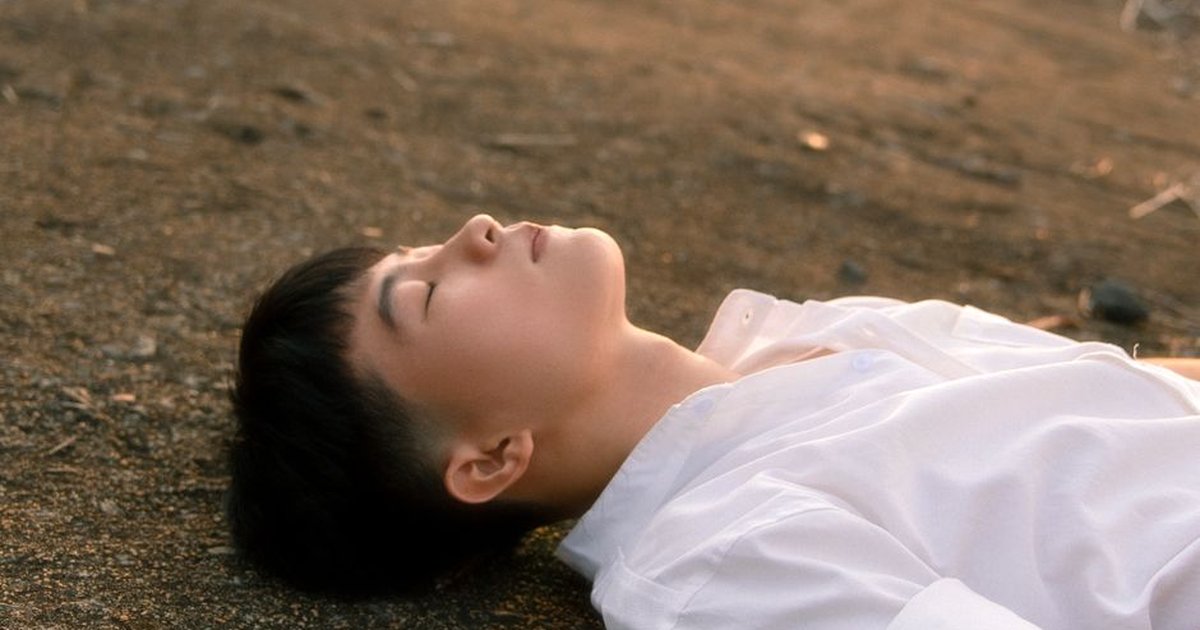A young man in a white shirt laying peacefully on the ground outdoors.