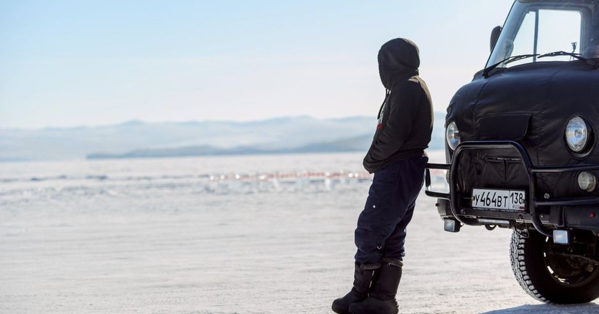 A person in winter clothing stands beside a van on a snowy landscape, under a clear sky.
