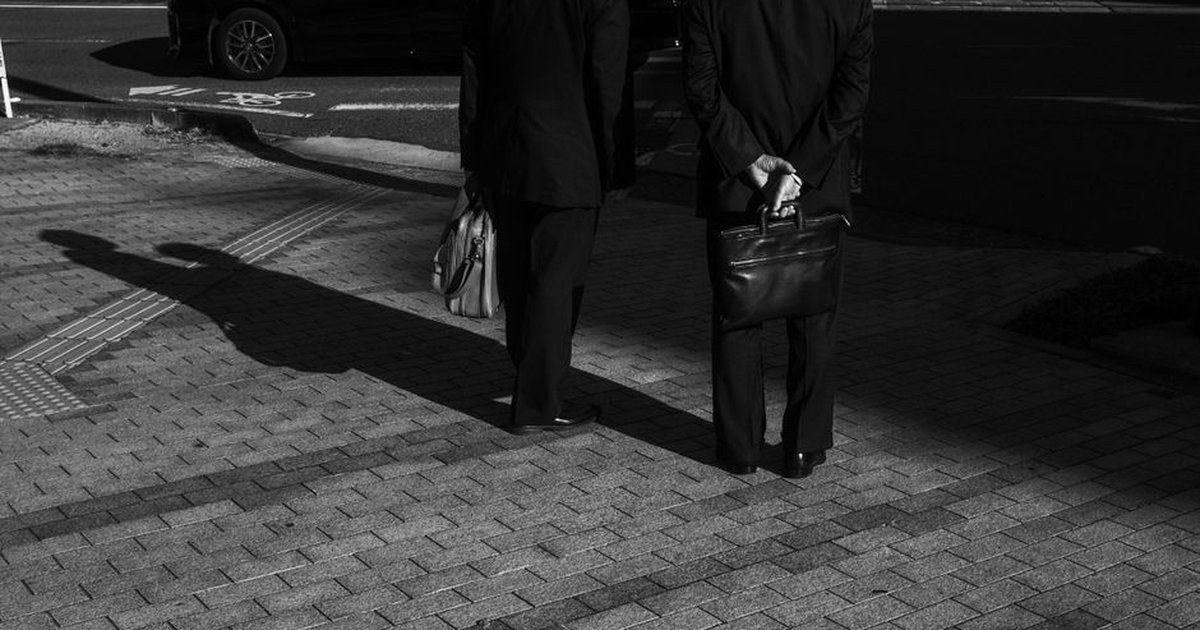 Men in suits with briefcases casting shadows on a sunlit city pavement.