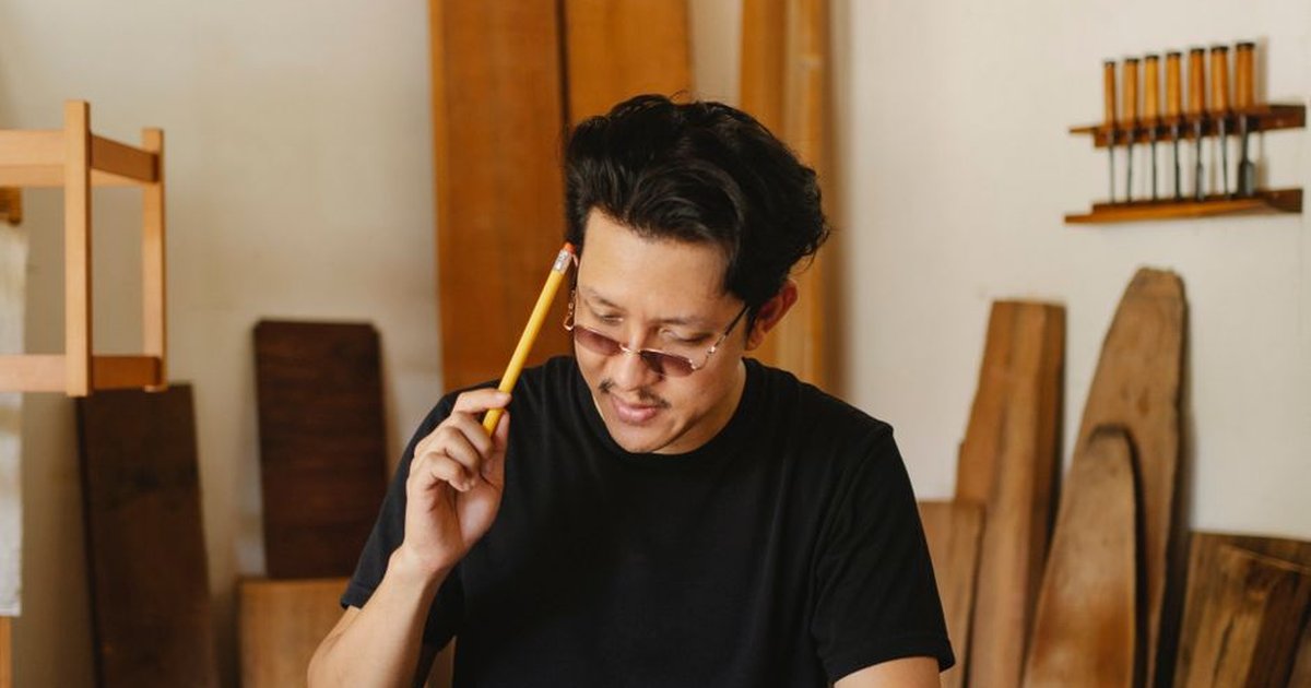 Charismatic ethnic male master in glasses holding pencil while creating new woodwork object in creative workplace