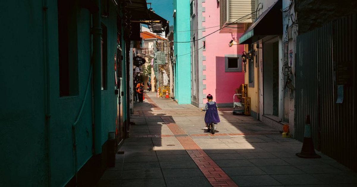 A child on a scooter explores a vibrant alleyway in Macau, filled with colorful buildings and warm sunlight.