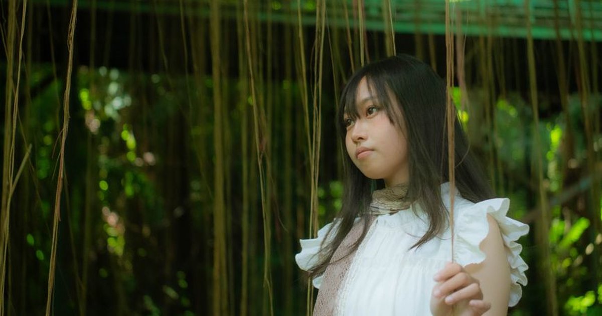 Portrait of a young woman in white dress surrounded by hanging vines in a lush forest setting.