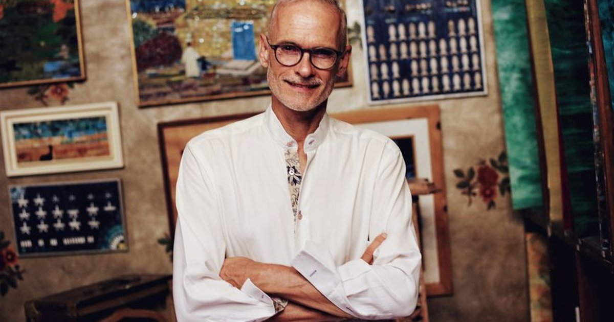 A senior man in glasses smiling while posing in an art gallery filled with paintings.
