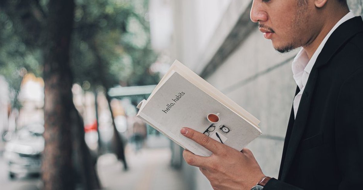 A man in a suit reading a book titled 'hello habits' on a city sidewalk.