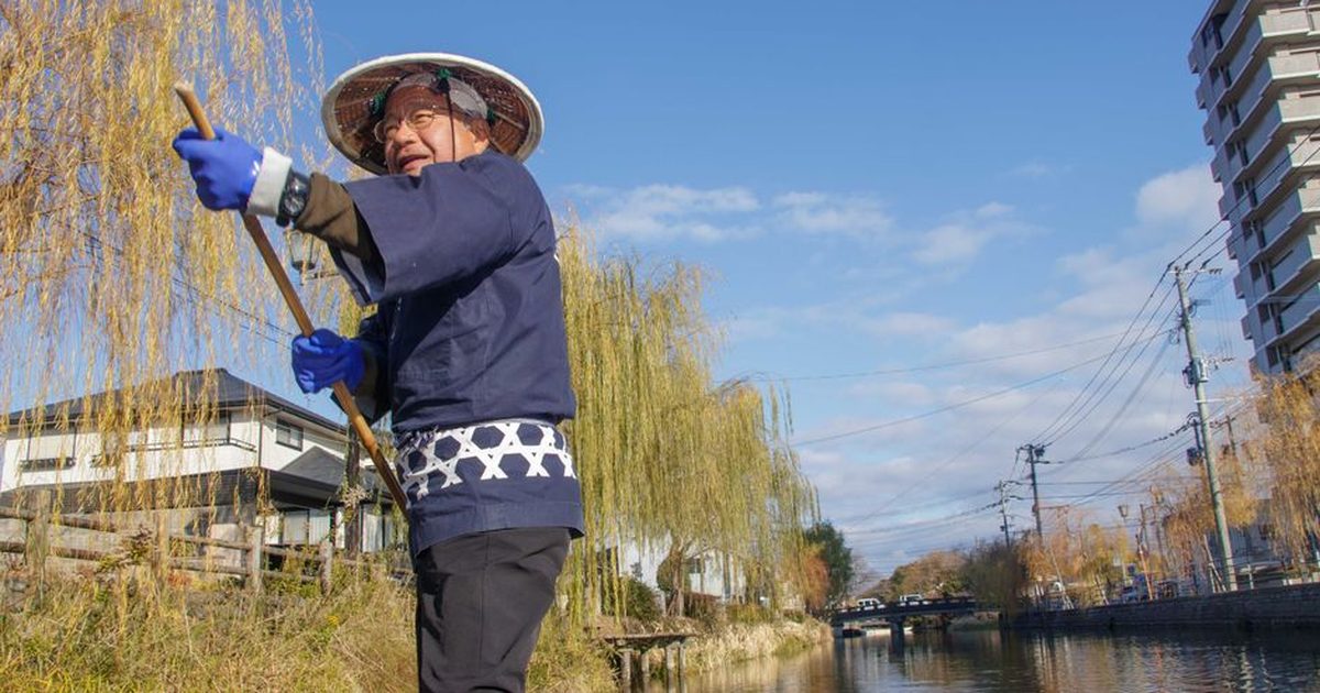 Elderly man wearing a traditional hat navigates a river on a sunny day with a lush backdrop.