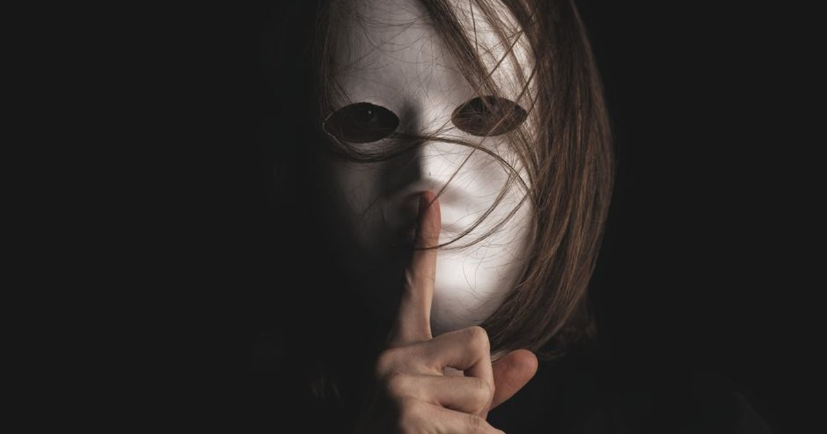 A person with a white mask makes a silence gesture, conveying mystery and secrecy.