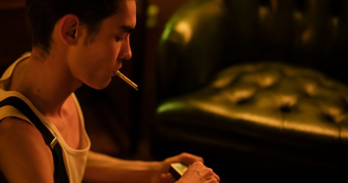 A young man sits indoors, smoking a cigarette in a dimly lit room.