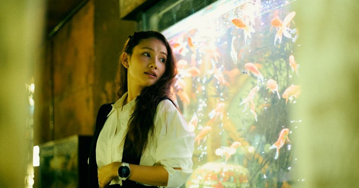 A woman gazes thoughtfully beside a vibrant aquarium filled with goldfish.
