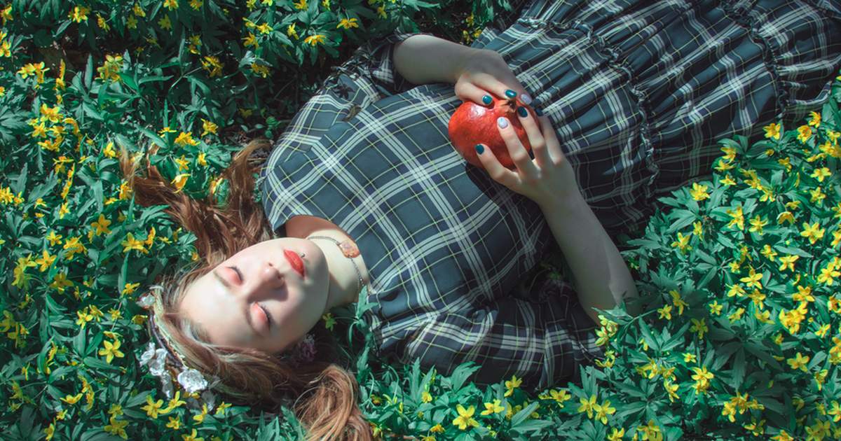 A woman lies in flowers holding a pomegranate, eyes closed, serene scene.