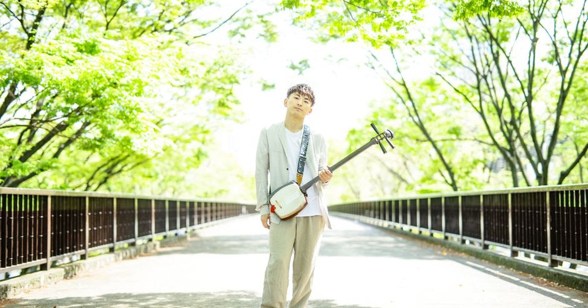An Asian man standing on a sunlit bridge, holding a shamisen, surrounded by vibrant green trees.
