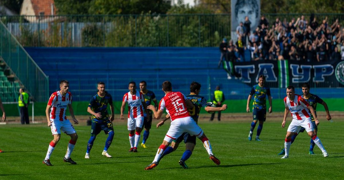 Intense soccer game with teams in action under bright sunlight, surrounded by cheering fans.