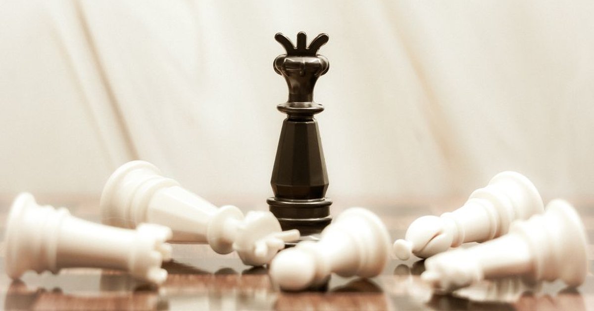 A black chess queen stands triumphant among toppled white pieces on a reflective surface.
