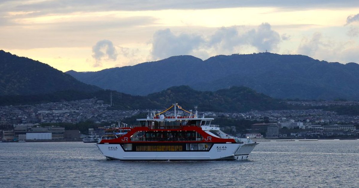 A JR West ferry sails near Hiroshima, Japan, at twilight, with scenic mountains in the background.
