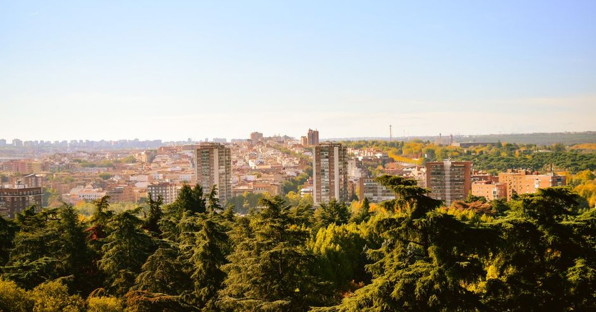 Panoramic cityscape view featuring lush green trees in the foreground and urban landscape in the background.