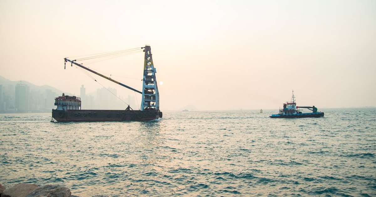 Tugboat guiding a crane barge on Kowloon harbor during a serene sunrise, overlooking Hong Kong's urban skyline.
