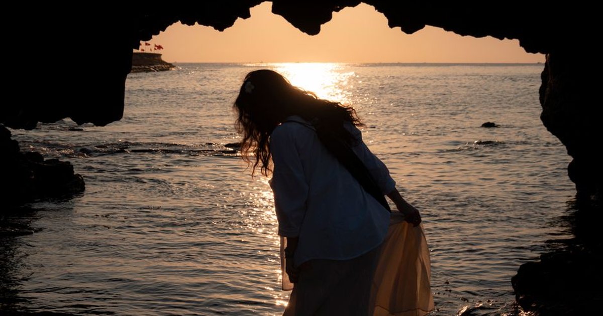 Silhouette of a woman in a cave, gazing at the serene sunset over the ocean. Peaceful and introspective.