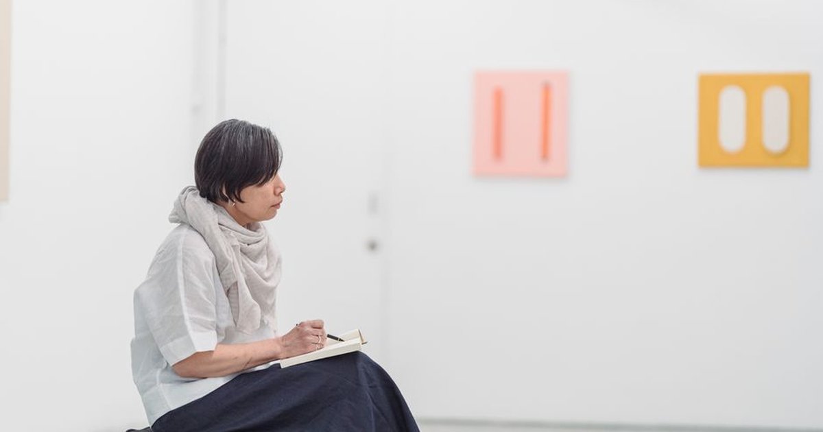 A woman thoughtfully sketches in a modern art gallery, surrounded by minimalist artwork.