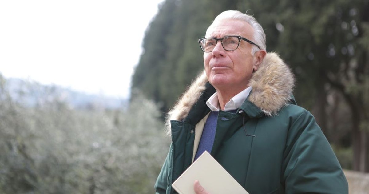 Senior man with glasses in a fur-collared jacket holding a book outdoors, looking thoughtful.