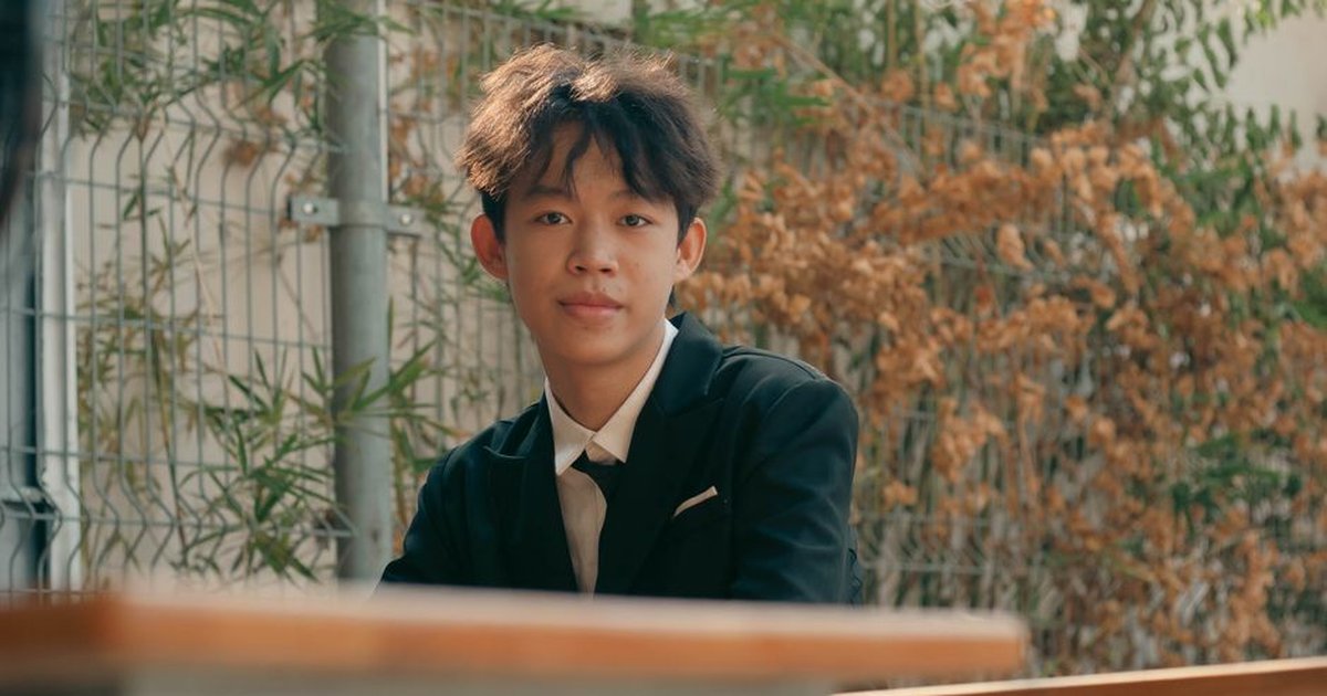 A young adult in a black suit sitting outdoors by a fence, exuding confidence.